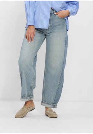 Vrouw draagt lichtblauwe brede jeans met omslag, beige instaploafers en een blauw gestreept overhemd met knopen, staand met één hand in de zak.