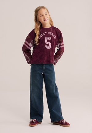 Jeune fille aux longs cheveux blonds portant un haut en dentelle bordeaux avec "Lucky Charm 5," jeans bleus larges, et des baskets bordeaux avec des rubans roses.