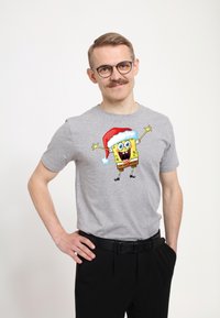 Šedé tričko s krátkým rukávem, na kterém je barevná grafika SpongeBoba v santa klobouku, nošené s černými kalhotami.