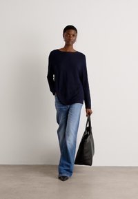 Marineblå langermet topp parret med lyseblå vide bukser. Modellen holder en svart tote-bag, mens hun står mot en nøytral bakgrunn.