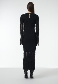 Schwarzes Kleid mit langen Ärmeln aus strukturiertem Stoff, Rückenteil mit Schlitz, geschlitzte Ärmel und goldene Akzente entlang der Seiten. Enganliegende Silhouette.