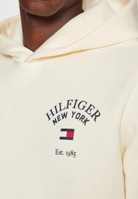 Felpa color crema realizzata in tessuto morbido. Presenta la scritta "HILFIGER NEW YORK" e la data di fondazione, con un piccolo logo in rosso, bianco e blu.