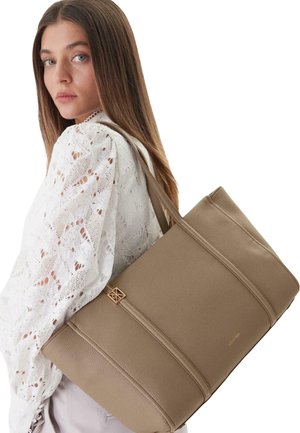 Giovane donna con blusa bianca in pizzo che porta una grande borsa tote in pelle taupe con finiture dorate sulla spalla.
