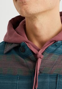 Blå och grön mönstrad skjortjacka med en vinröd huva. Tyget har en strukturerad finish och har en knuten dragsko.