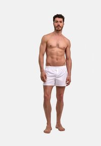Witte katoenen shorts met een elastische tailleband, gedragen door een man met een slank postuur die blootsvoets staat tegen een effen grijze achtergrond.