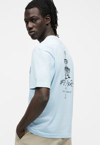 Νέος άνδρας με σκούρο δέρμα και κοτσίδες που φορά ανοιχτά μπλε μπλουζάκι με μαύρη εικόνα κοκτέιλ στο πίσω μέρος και μπεζ παντελόνι.