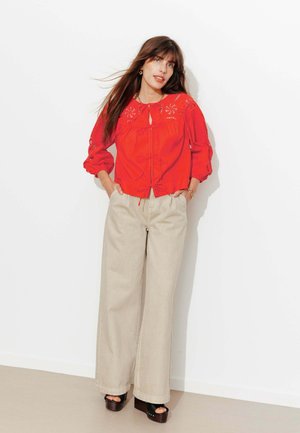 Femme aux longs cheveux bruns portant un chemisier rouge brodé, un pantalon beige à jambes larges et des sandales compensées noires, debout contre un mur blanc.