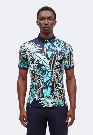 Homme portant un polo à manches courtes avec des motifs de feuilles tropicales bleu, sarcelle, blanc et marron et un pantalon foncé sur un fond uni.