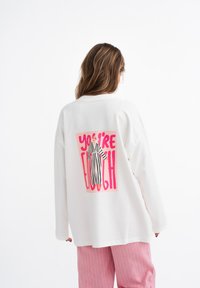 Witte oversized sweatshirt met een grafische print op de achterkant met de zin "JIJ BENT GENOEG" in roze en een gestreepte karakterillustratie.