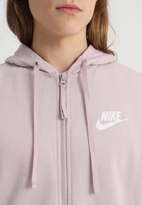 Sudadera con cremallera Nike de color rosa claro, con una textura suave, cordones ajustables y un logotipo blanco prominente en el lado izquierdo del pecho.