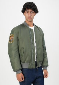 Groene bomberjack met grijze geribde kraag en manchetten, voorrits, zijzakken en een tijgerpatch op de linkermouw.