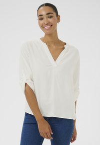 Blusa leggera e bianca in tessuto con scollatura a V e maniche arrotolabili. La texture è leggermente patterned, offrendo una vestibilità comoda, abbinata a jeans blu.
