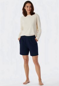 Vrouw staat blootsvoets in een crèmekleurig langarmshirt en donkerblauwe elastische shorts met haar handen in de zakken.