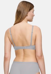 Ženska z dolgimi svetlo rjavimi lasmi, oblečena v sivo nedrček in usklajeno spodnje perilo, prikazana od zadaj na enobarvni beli podlagi.