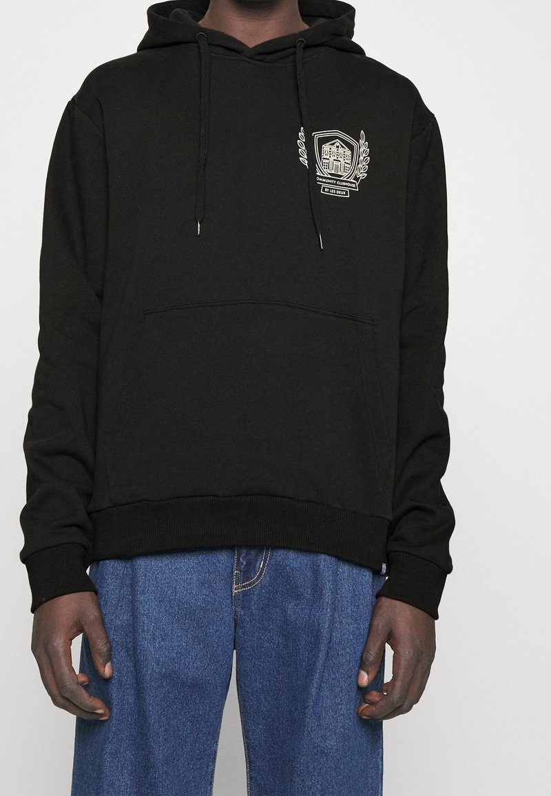 Sweat à capuche noir avec poche avant et emblème blanc sur le côté gauche de la poitrine, porté avec un jean en denim bleu sur une personne à la peau foncée.