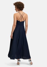 Granatowa długa sukienka maxi z krzyżującymi się ramiączkami na plecach, dopasowaną talią i zwiewnym fasonem spódnicy. Posiada gładką fakturę i subtelny połysk. Srebrne szpilki.