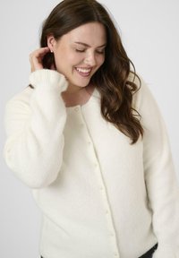 Smiling woman with long brown hair touches her ear, wearing a white buttoned cardigan with pearl-like buttons against a plain background.