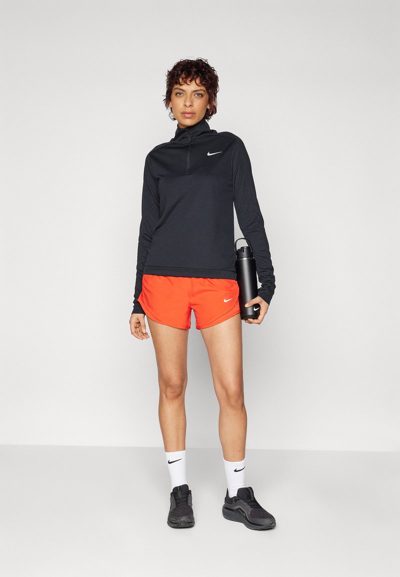 Top negro de manga larga con cuello con cremallera, pantalones cortos naranjas, zapatillas negras y una botella de agua. Presenta logotipos de Nike en la parte superior y en los pantalones cortos.