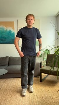 Bărbat îmbrăcat cu un tricou negru și blugi stă în living, lângă o canapea gri, un fotoliu verde, plante și artă abstractă pe perete.