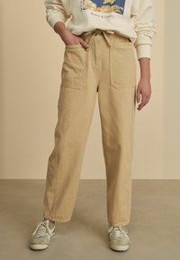 Beige ribfluwelen broek met verticale strepen, voorzien van grote voorzakken en een losse pasvorm. Gecombineerd met grijze sneakers.