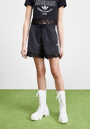 Personne portant un short noir Adidas avec bordure en dentelle, un crop top noir Adidas et des bottes blanches montantes lacées jusqu’au genou, debout sur un sol vert.
