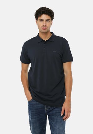 Uomo che indossa una polo blu scuro e jeans blu, in piedi con una mano in tasca contro uno sfondo bianco neutro.