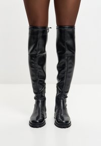 Botas de cuero negras hasta la rodilla con una textura suave, detalle de cremallera en el lateral y suelas de goma resistentes para mayor tracción.