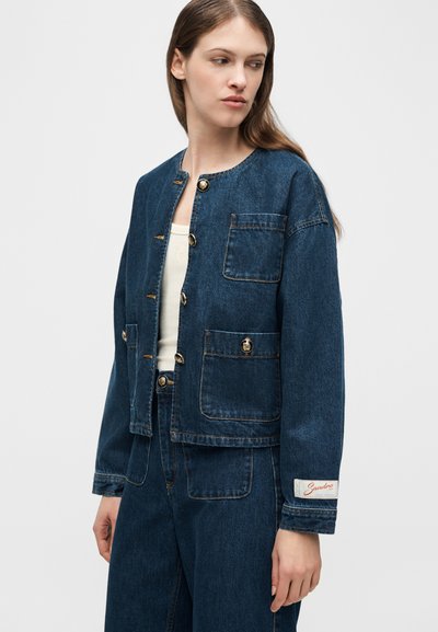 Γυναίκα που φοράει ένα σκούρο μπλε τζιν μπουφάν με χρυσά κουμπιά, τσέπες με επάλειψη και τζιν παντελόνι με ψηλή μέση που ταιριάζει, κοιτάζοντας προς τα πλάγια.