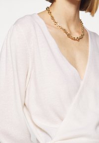 Femme portant un pull cache-cœur blanc à col en V et un gros collier chaîne en or, montrée du cou jusqu'au milieu du torse.