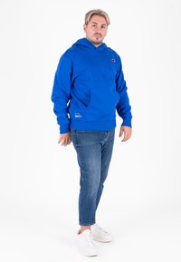 Felpa blu con tasca frontale e cappuccio, realizzata in un tessuto morbido. Abbinata a jeans azzurri e sneakers bianche.