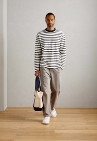 Man walking indoors wearing a white and black striped long-sleeve shirt, beige pants, white shoes, carrying a black and beige tote bag.