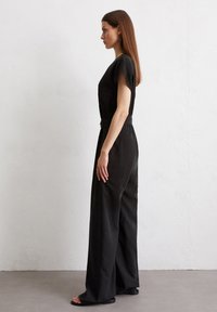 Zwarte jumpsuit met korte mouwen, wijde broekspijpen en een strikceintuur in de taille. Soepele textuur, minimale naden en een effen kleur.