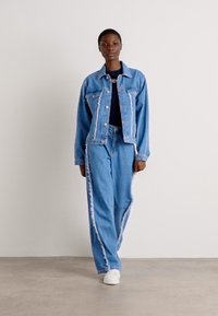 Jeansjacke und Hosen-Set in Hellblau mit ausgefransten Kanten und seitlichen Akzenten. Die Jacke verfügt über Fronttaschen und einen Knopfverschluss.