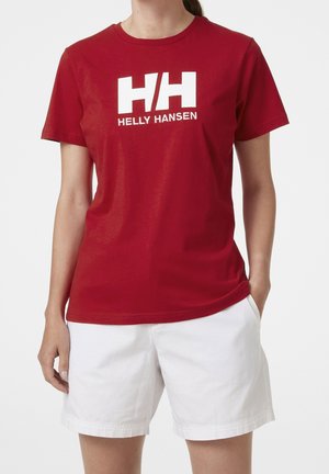 Persoon draagt een rood Helly Hansen T-shirt met wit logo en tekst, gecombineerd met witte shorts, staand met één hand in de zak.