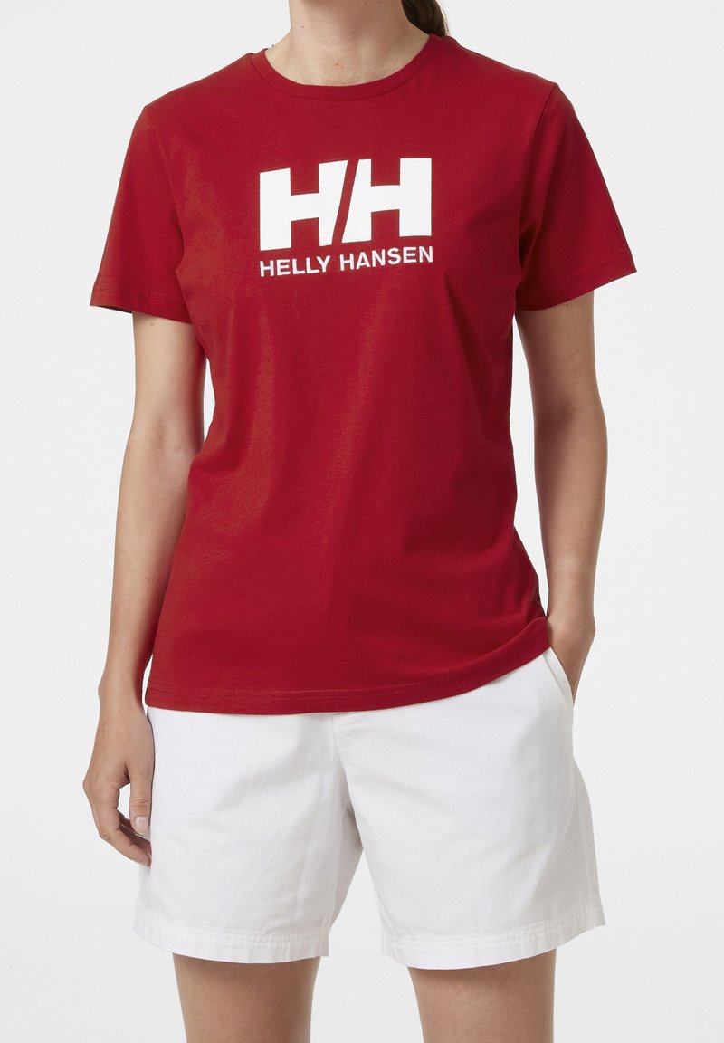 Personne portant un t-shirt rouge Helly Hansen avec logo blanc et texte, associé à un short blanc, debout avec une main dans la poche.