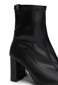 Botas de cuero negro hasta el tobillo con una textura suave. Presentan un tacón cuadrado y un cierre de cremallera en la parte superior. Diseño elegante y minimalista.