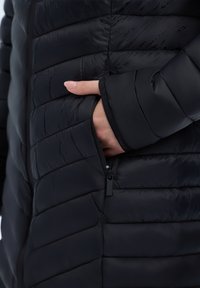 Musta puhallettu takki vaakasuoralla tikkauksella, jossa on vetoketjullinen tasku ja pehmeä sisävuori hihansuissa, korostaen sen rakennetta.