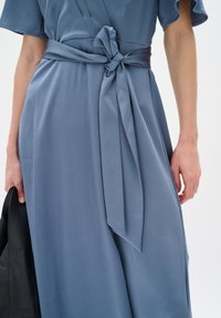 Helles blaues Kleid aus strukturiertem Stoff, mit einem taillierten Gürtel und einer Schleife. Das Design umfasst kurze, ausgestellte Ärmel und eine fließende Silhouette.