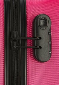 Valise rigide rose avec une surface texturée, une fermeture éclair noire renforcée et un mécanisme de verrouillage à combinaison sur le côté.
