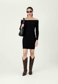 Vestido de malha preto, com ombros à mostra e textura canelada, usado com botas de cowboy castanhas e uma bolsa de ombro preta. Mulher com cabelo trançado e óculos de sol.