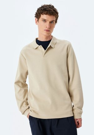 Beige poloshirt met lange mouwen, een kraag, een placket met twee knopen en een gladde textuur. Draagt over een marineblauwe basislaag.