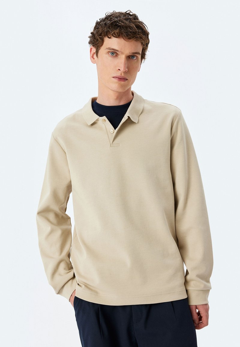 Beige pitkähihaisessa polo-paidassa on kauluspääntie, kaksi napin lista ja sileä pinta. Tätä käytetään tummansinisellä peruskerroksella.