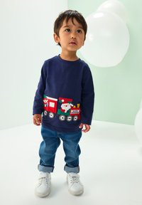 Marinblå långärmad skjorta med filtapplicering av ett färgglatt tåg med jultomten, röda och gröna detaljer, kombinerad med blå denimjeans och vita sneakers.