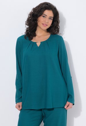 Kvinde med krøllet mørkt hår iført teal langærmet top med plisseret halsudskæringsdetalje og matchende bukser, stående mod en hvid baggrund.