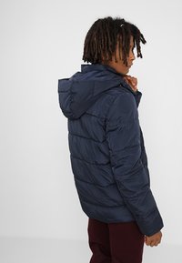 Marineblå dunjakke med hætte, quiltet design, glat tekstur og synlige stikninger. Har en tætsiddende pasform og manchetter på ærmerne.