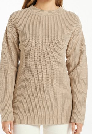 Pull en maille côtelée beige à manches longues et col rond porté par une personne avec un pantalon clair, visible des épaules jusqu’aux hanches.