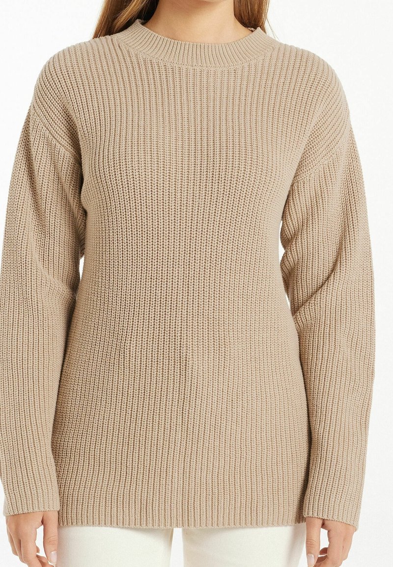 Pull en maille côtelée beige à manches longues et col rond porté par une personne avec un pantalon clair, visible des épaules jusqu’aux hanches.