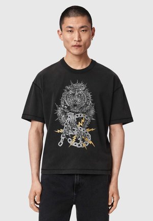 Homme portant un t-shirt noir avec un graphisme d’une tête de tigre épineuse, une griffe enchaînée et des éclairs jaunes sur le devant, debout devant un fond uni.