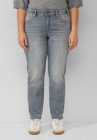 Osoba w szarych wyblakłych dżinsach i białych trampkach stoi na jednolitym jasnoszarym tle.