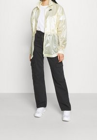 Ljust gul, genomskinlig vindjacka med dragkedja och elastiska manschetter, kombinerad med svarta cargo-byxor och vita sneakers.